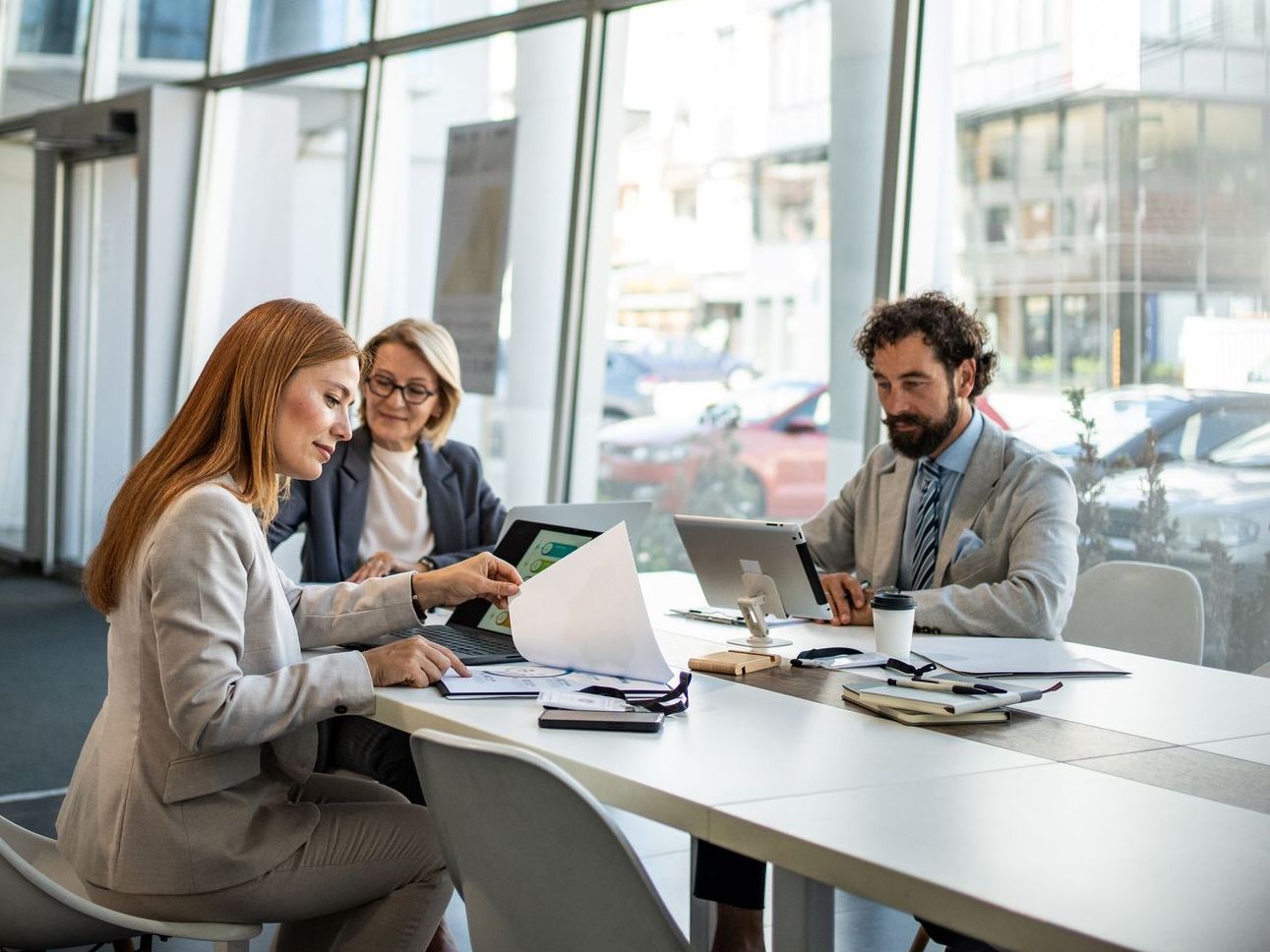 Three business professionals ina modern office meeting around a white table, reviewing documents and using laptops.