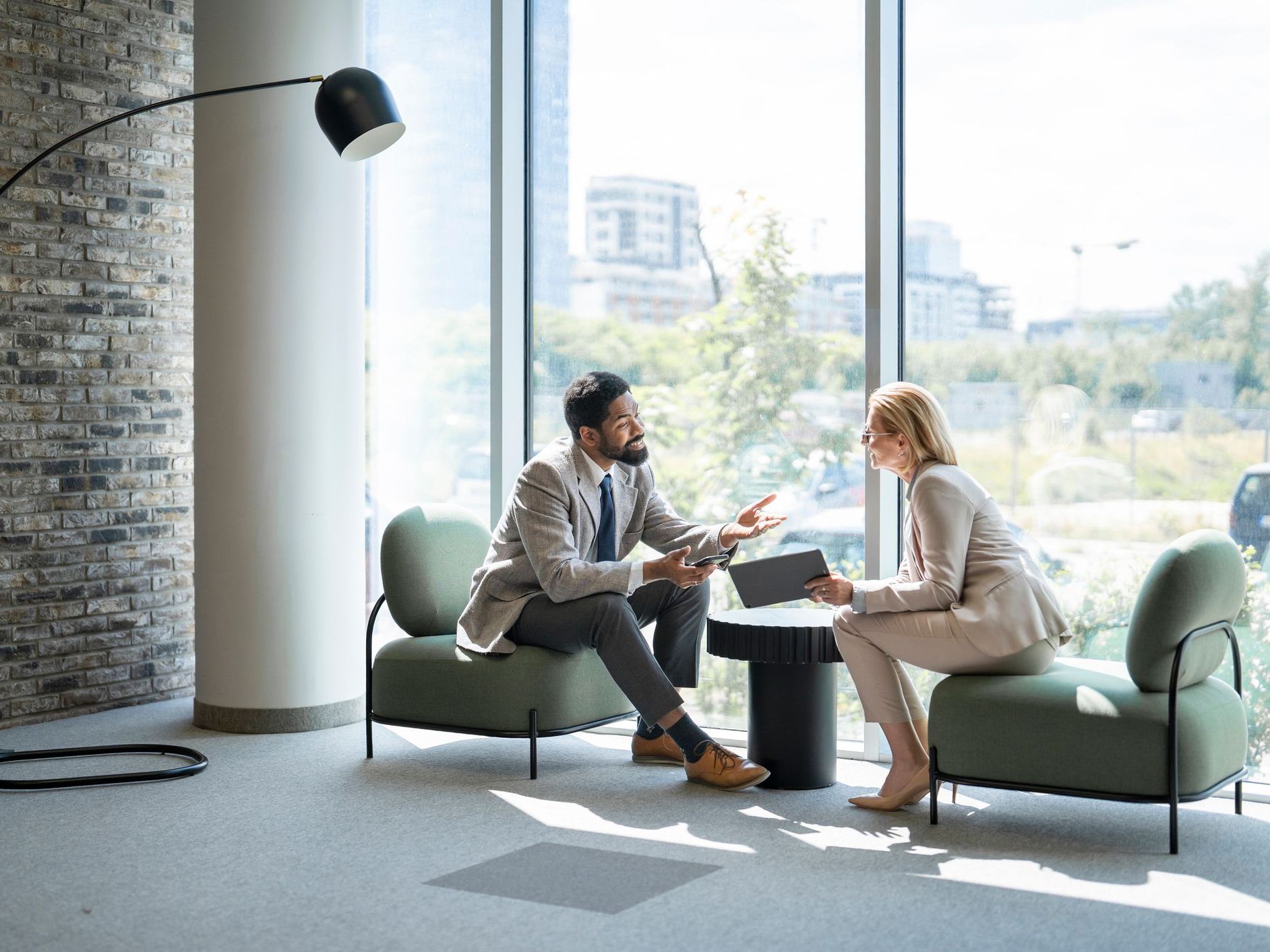 Man and woman in business attire sit in green chairs by a window, talking while holding a phone and tablet.