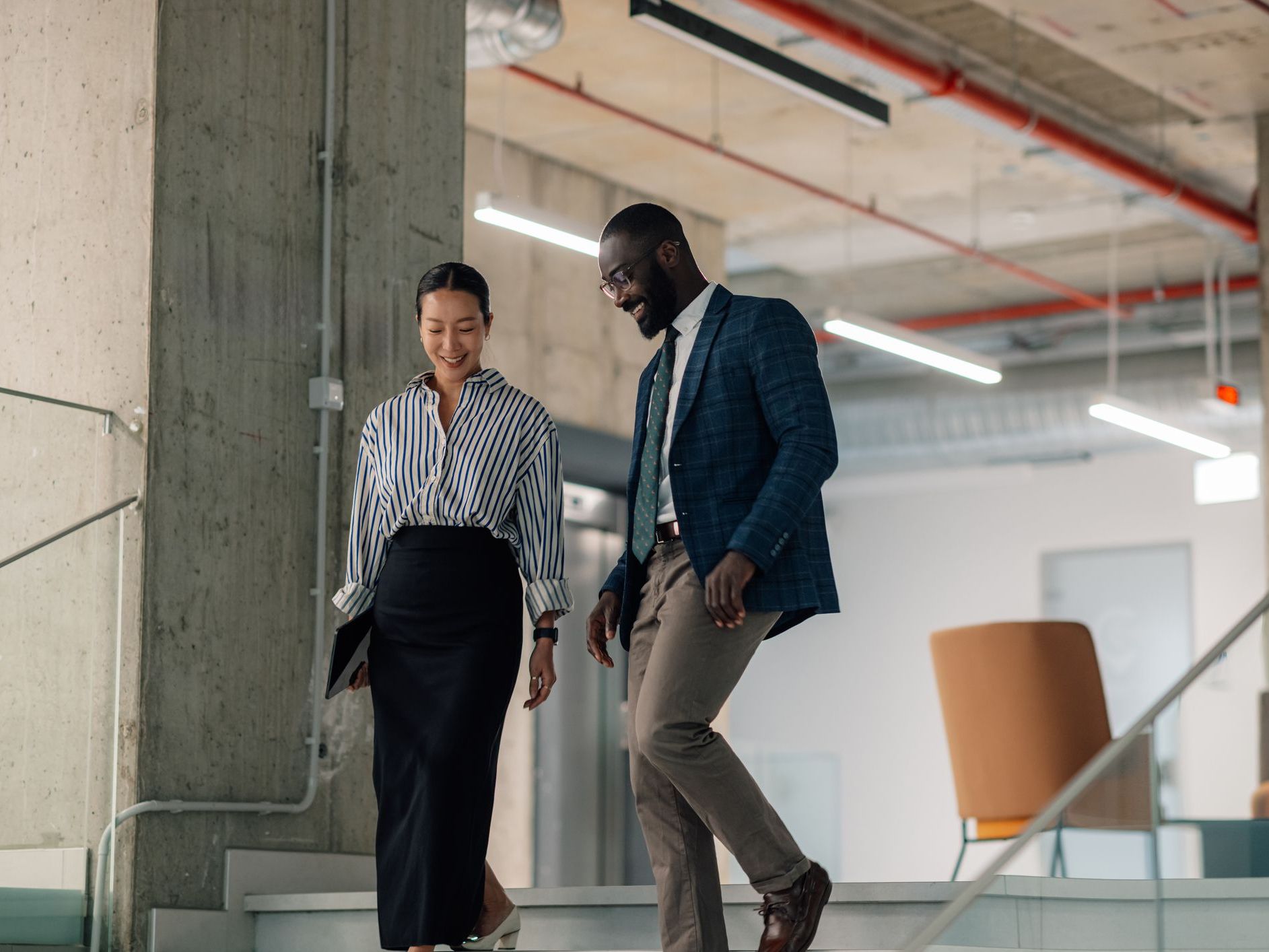 Two business professionals, a woman in a striped shirt and a man in a blue blazer, walk down stairs in a modern office.