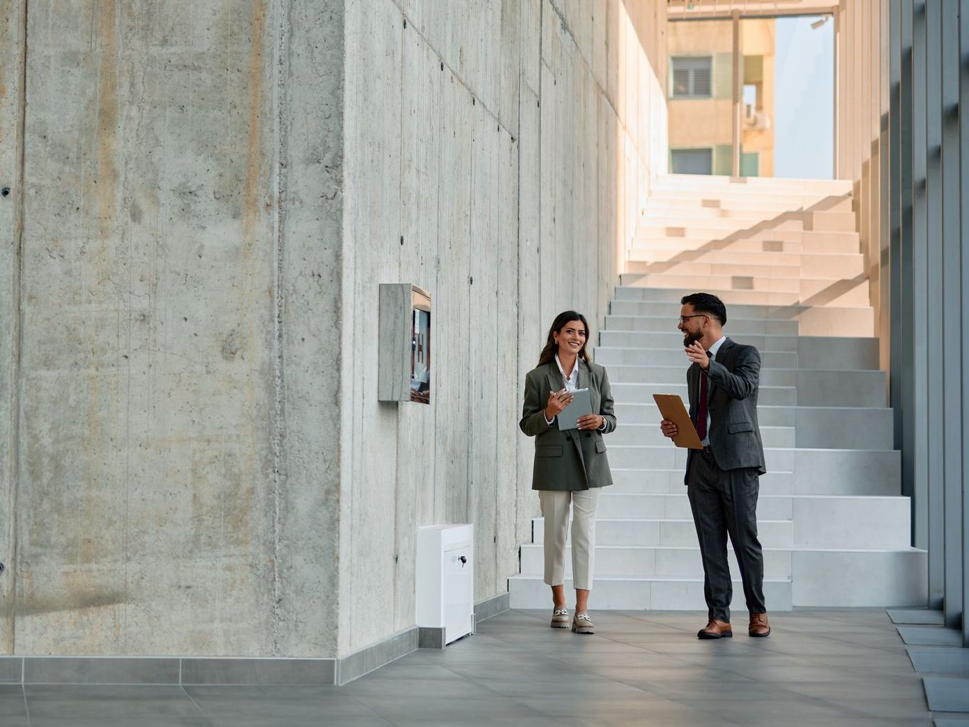 Man and woman in businessattire discuss near a concrete wall with a mounted device, holding clipboards, with stairs.