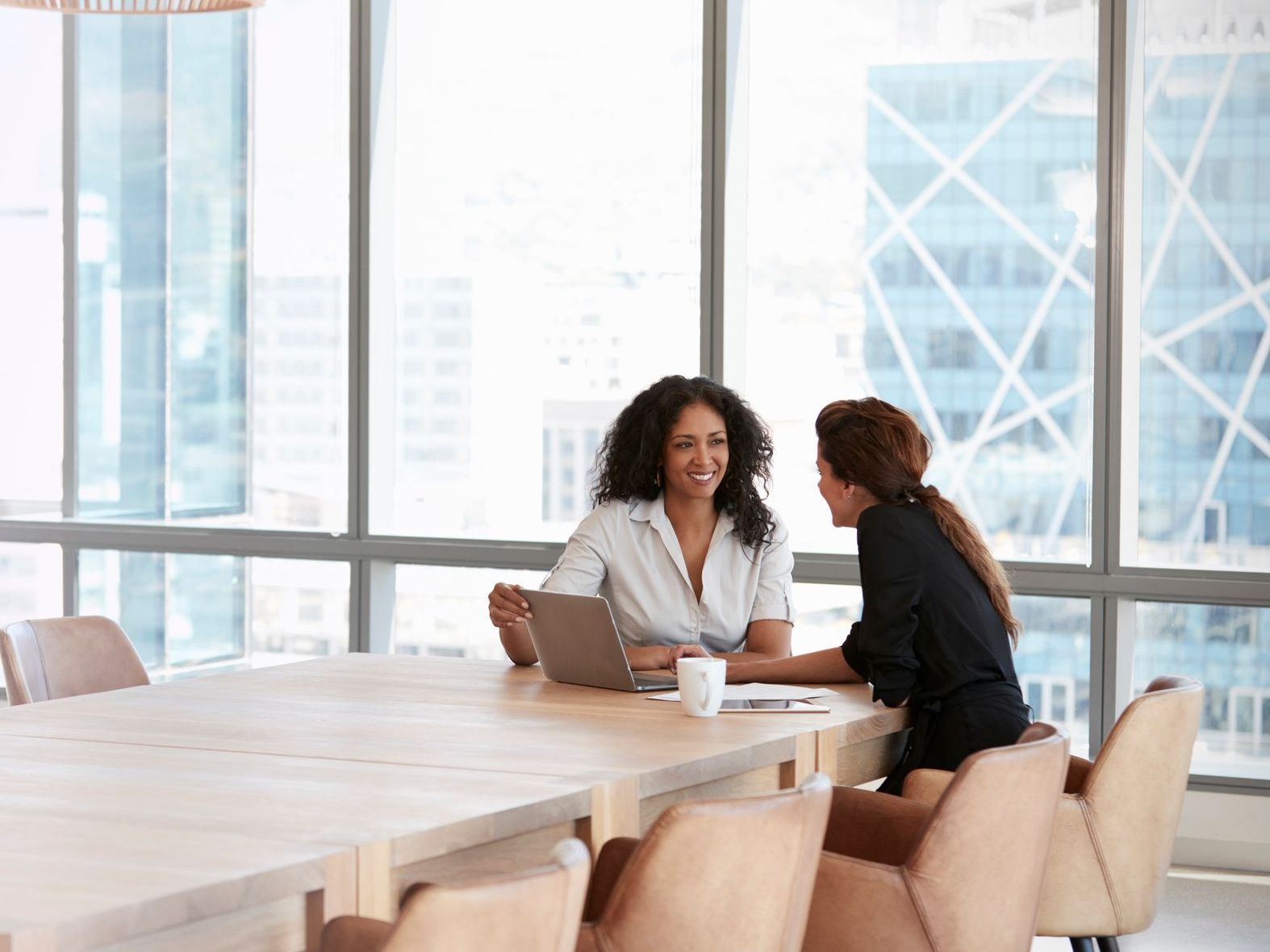 Two women meet at a wooden conference table with a laptop, smiling, in a bright office with city view windows.