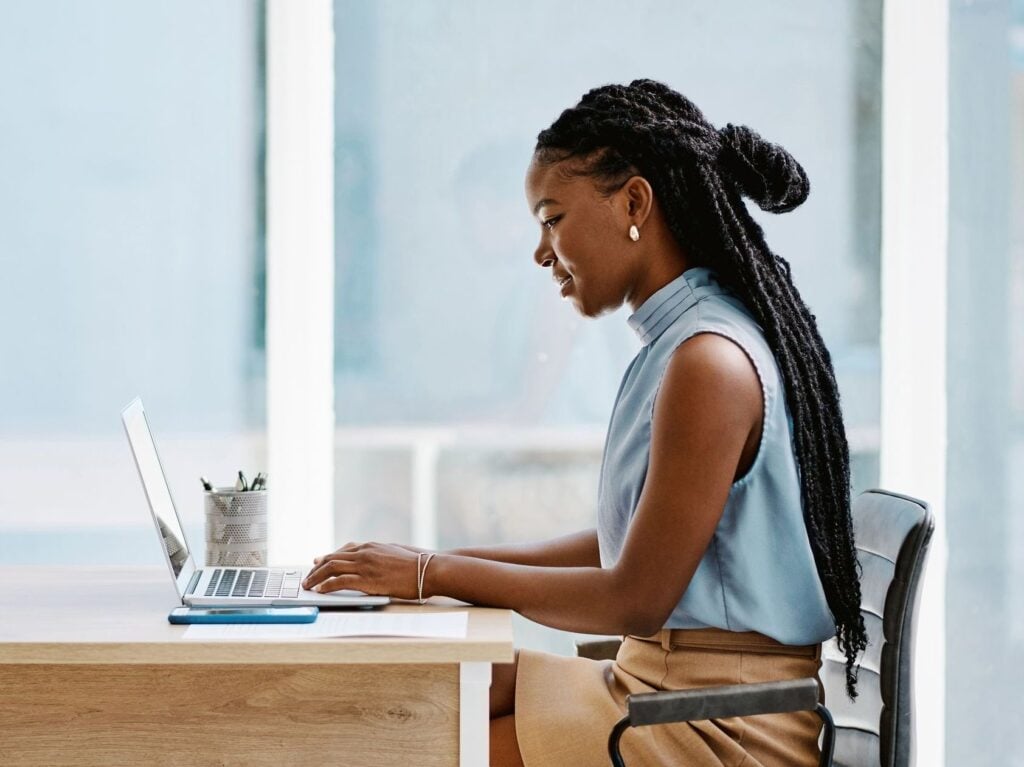 Woman with braided hair working on a laptop at a desk, with a window in the background.